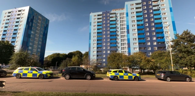 Two tower blocks of about 15 storeys. They are coloured light and dark blue on the outside. A line of cars and police cars is parked on the road in the foreground.