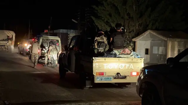 Trucks, a motorbike and cars carrying armed men are driving along a dark road in a convoy