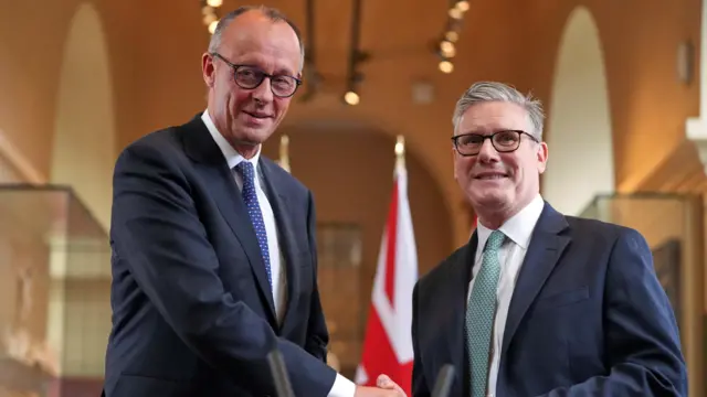 Merz and Starmer stand inside the V&A building smiling and the camera shaking hands.