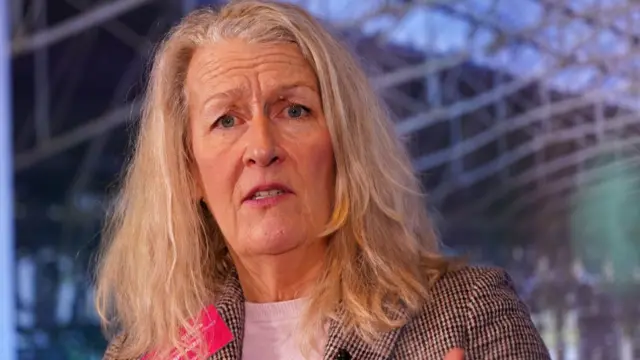 A woman with blonde hair and a pink badge on her blazer speaks as she looks down towards the camera which is slightly below her