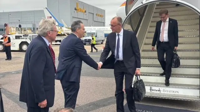 German chancellor shakes hand with ambassador at the bottom of a flight of aeroplane stairs. A man walks down the final steps carrying a black bag.