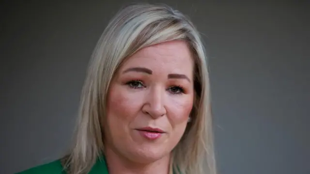 Headshot of a woman with a blonde bob speaking. There's a plain grey background behind her