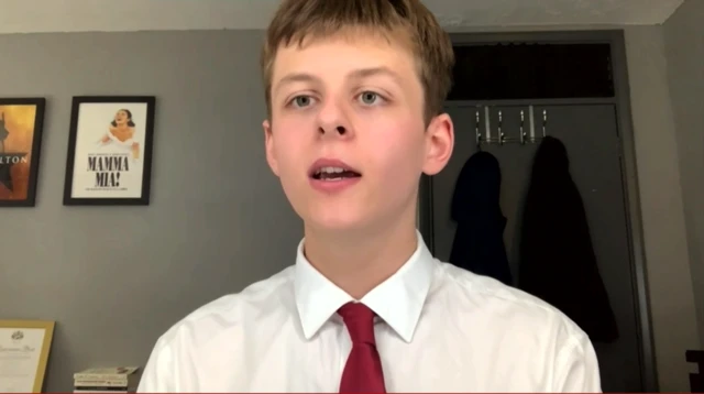 Young adult in white shirt and red tie speaking from inside a room with grey walls, a poster of the musical Hamilton on the wall behind him, another one for the musical Mamma Mia! next to it