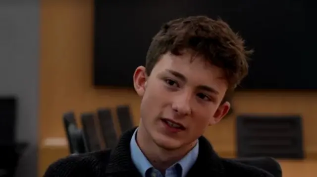 Headshot of a boy who sits and cocks his head while he talks. He is in a meeting room with a wooden table and wall, and black chairs