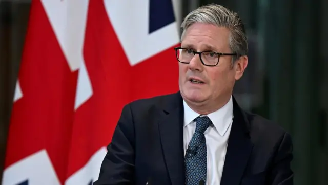 Headshot of Starmer who sits and speaks, wearing a small microphone clipped to his tie. Behind him you can see a large British flag hanging.