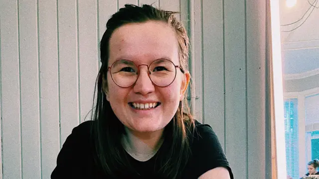 A young brown-haired woman with large round glasses smiles to the camera while sitting down inside what appears to be a restaurant. She's wearing a black short-sleeved t-shirt