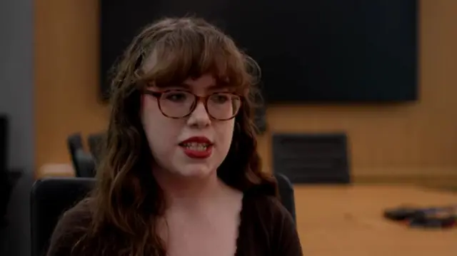 Headshot of a girl who is speaking look past the camera. She is in a meeting room with a wooden table and wall, and black chairs