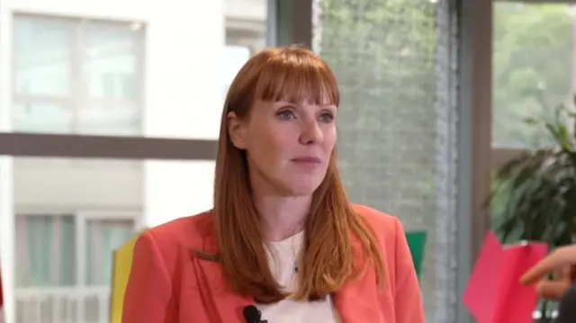 Headshot of Rayner who looks at someone in front of her who is off camera. She stands in front of windows