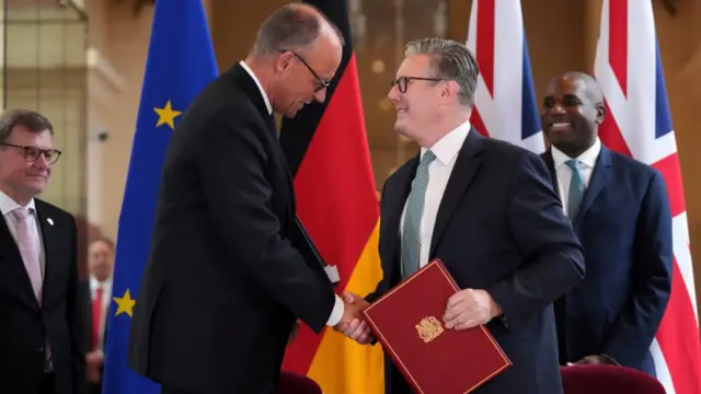 Starmer and Merz shake hands as Starmer holds a red folder containing the treaty. On either side of them, Lammy and Wadephul watch on