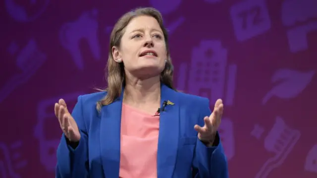 Helen Maguire gestures with her hands wearing a blue jacket and pink top.