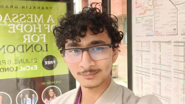 Selfie of Aqdas who wears glasses and has curly hair. He stands in a bus stop with the map behind him and a poster behind him