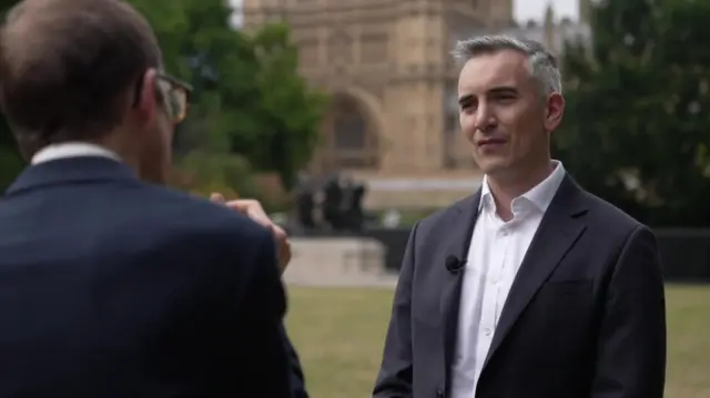 Back of journalist in black suit (L) as he speaks to a man in black suit, white shirt on a grassy patch, Westminster Cathedral partially visible in the background