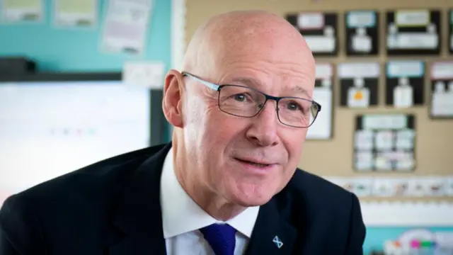 Headshot of Swinney who looks forward past the camera. He is sitting in a classroom, the background is blurry