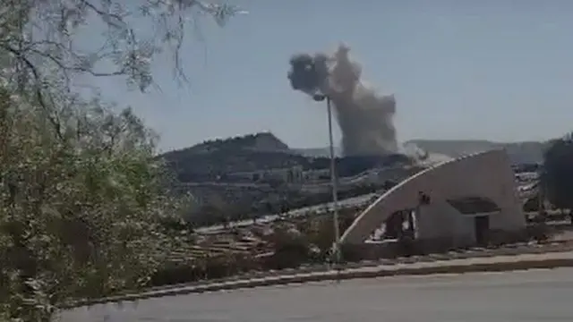 A screenshot showing a road. In the background smoke can be seen rising from a hillside near the presidential palace in Damascus