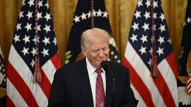 Donald Trump smiles while speaking into a microphone at a lectern during an event in the White House's East Room with American flags behind him.