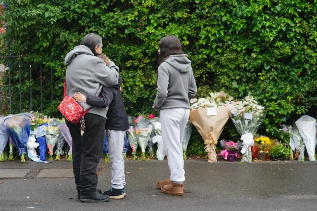 A woman wearing a grey hoodie and a red cross-body bag hugs a small boy wearing a black jacket and grey joggers. A young woman stands next to them. They are all looking at floral tributes lined up along the street.