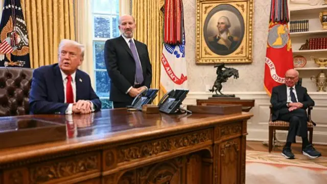 Donald Trump sits at his desk in the oval office, with Secretary of Commerce Howard Lutnick standing near him and Rupert Murdoch sitting on a chair nearby