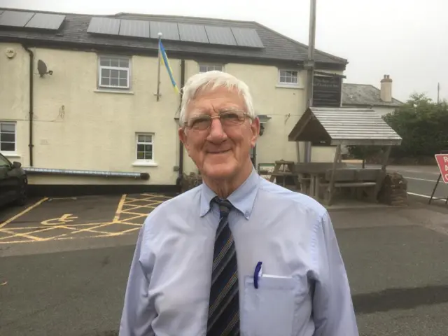 Eric Norman, who wears a light blue shirt and a dark striped tie, stands in front of the pub. He is smiling.
