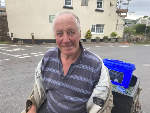 Roger, who wears an open light-coloured jacket and a striped top, looks at the camera. He is standing in the middle of the street.