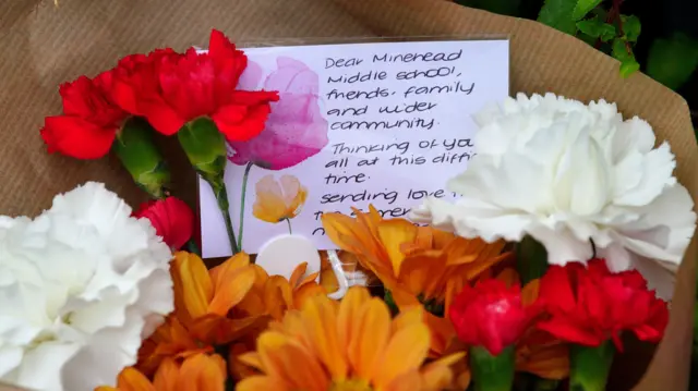 A bunch of orange, red and white flowers with a note saying: "Dear Minehead Middle School, friends, family and wider community. Thinking of you all at this difficult time. Sending love...". The rest of the message is hidden behind flowers.
