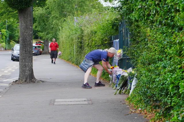 A person with long grey hair places a bunch of flowers at the school gate.