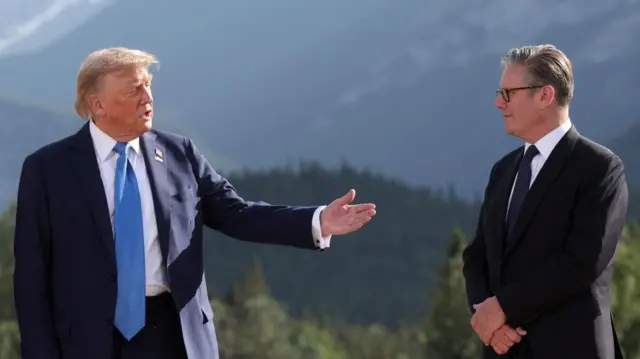 Donald Trump getures with his hand towards Keir Starmer, standing opposite him with arms folded. Both men in suits in front of a mountainous forest