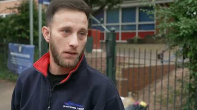 Waren Roland stood outside the school gates. He is visibly upset and looking to one side.