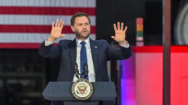 JD Vance stood behind podium, both hands in the air with palms facing out towards camera, US flag behind him.