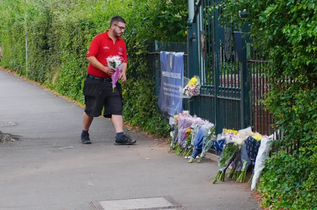 A man in a red work polo and long black shorts approaches the gates of Minehead Middle School with a bunch of flowers. A number of flowers are already in place.