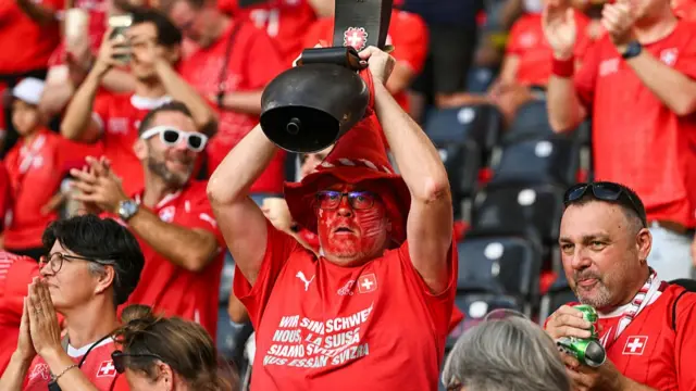 A supporter with a face painted with the colour of the Swiss flag holds a giant bell