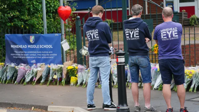 Three men facing floral tributes outside Minehead Middle School. They are wearing hoodies and t-shirts saying Minehead Eye Youth Services on the back. There is a long line of flowers on the floor.