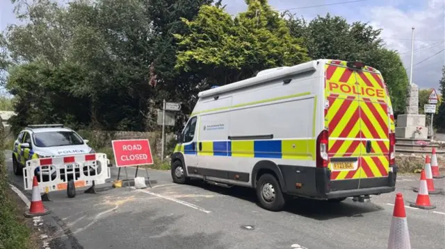 A police van facing away from the camera and a police car facing it behind a white plastic barrier. There is a red road closed sign in between them and cones behind the van.