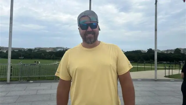 James from Tennessee smiles at the Camera. He is wearing a backwards facing baseball cap, sunglasses and a yellow shirt