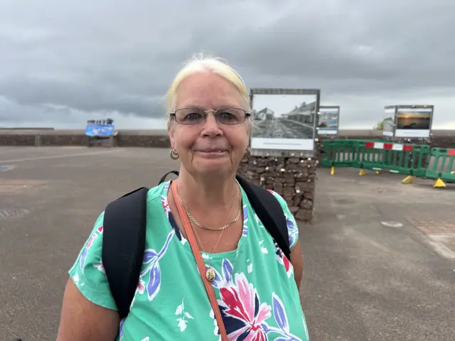 Cindy looks at the camera with a small smile on her face. She is shown in a large tarmac area in front of the seafront. She has very light blonde hair, and wears a green t-shirt with large pink and purple flowers on it.