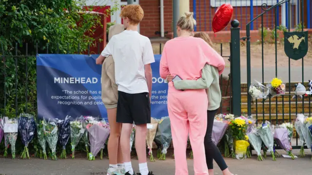 People comfort each other as they look at the floral tributes at the entrance to the Minehead Middle School in Minehead Somerset. They are facing away from the camera. A woman in a pink tracksuit and a girl are hugging. A boy in a white top with red hair is hugging another younger girl.