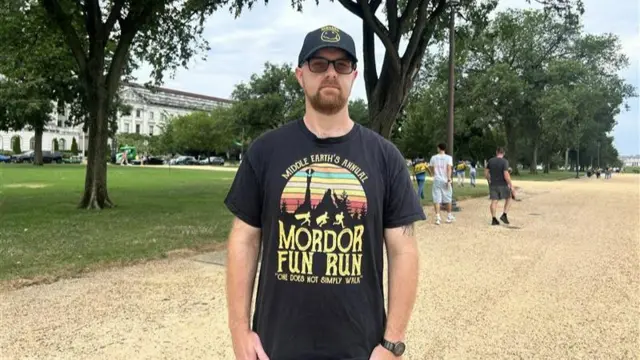 Man in a black t-shirt and baseball cap faces the camera in front of the National Mall.
