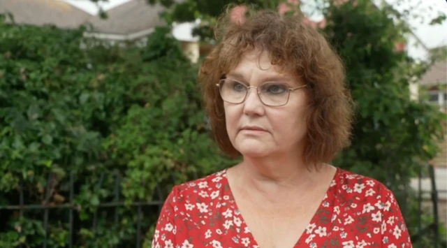 Trish Beckett stood outside the school in Minehead. There is a metal fence and shrubbery behind her. She has short brown hair which is curly. She is wearing a red top with green and white flowers on it. She also has metal-rimmed glasses on.