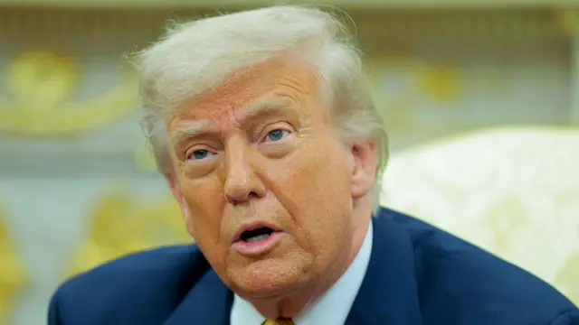 Close up of Donald Trump in blue suit jacket, white shirt and top of a gold tie visible. He's sitting in the Oval Office, the back of a yellow and white brocade chair visible behind him in the blurred background