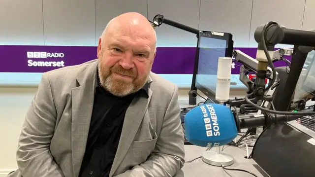 Bill Revans in BBC Somerset Radio's studio, he is wearing a grey suit jacket.