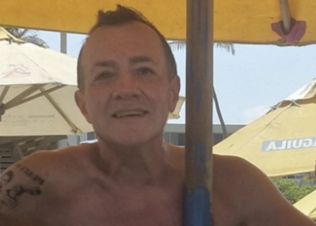 An image of a man with a tattoo on his right arm, smiling at a camera as he sits on a beach underneath a beach umbrella.