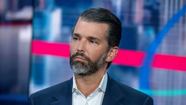 Headshot of Trump Jr who has a grey and black beard and slicked back black hair, as he sits and looks off to the side