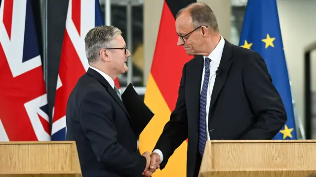 Starmer shakes hands with Merz in front of two wooden lecterns. Behind them are two UK flags, one Germany flag, and one European Union flag. Both men are wearing black suits.