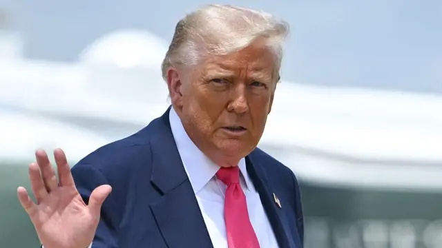 Donald Trump in dark blue suit, white shirt and red tie waves with his right hand with a serious expression on his face