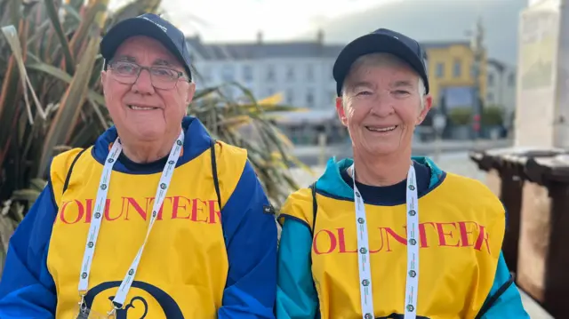 Mervyn and Sandra Best both wearing yellow vests and blue jackets underneath. Both are wearing navy hats and white lanyards. Mervyn is wearing glasses.