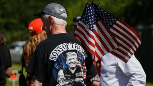 Elderly man in black t-shirt with an image of Trump's head superimposed on a biker's body, the writing Sons of Trump in a white banner at the top of the image. The man is holding two US flags, a woman with her back to the camera wearing a red cap stands ahead of him to his right