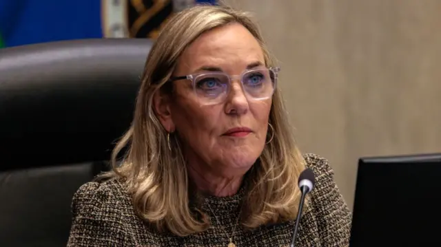 Kathryn Barger sits in front of a microphone. She has short blonde hair and blue eyes. She is wearing square-shaped glasses with a transparent rim.
