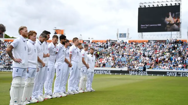 England players line up to pay tribute to Syd Lawrence before the England v India Test match at Headingly. Syd is pictured on a large screen in the background and the grandstands are full of spectators. The players are wearing black armbands