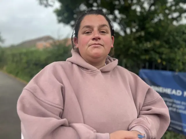 Leanna, who is wearing a light pink hoodie and has dark brown hair, looks at the camera with a serious expression. She is standing on the pavement outside the school.