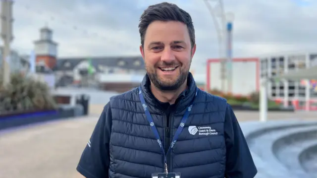 Jonny McCarron, he is looking into the camera and smiling. He has dark hair and is wearing a navy gilet that is council branded. He has a navy polo underneath and a white and navy lanyard,