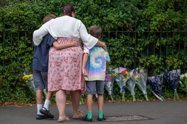 Mum cuddles two sons while stood near floral tributes